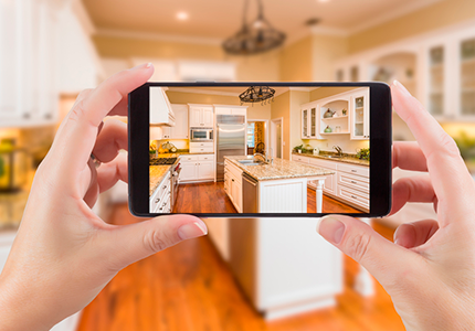 Simulador_de_ambientes_online-1Fotografia de mãos segurando um celular que mostra uma cozinha com piso de madeira e móveis brancos.