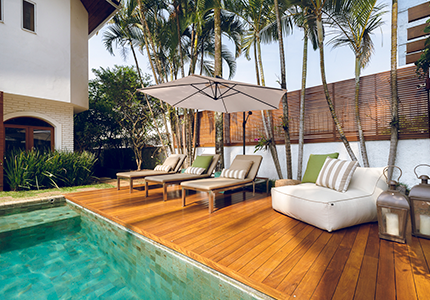 Fotografia de uma área externa com deck de madeira, piscina, almofadas e poltonas.