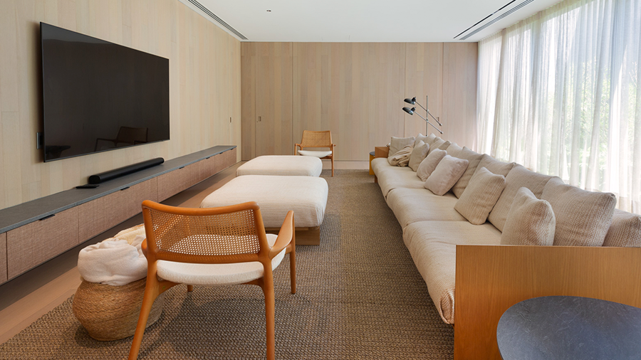 Imagem de uma sala com a parede revestida com a madeira clara tauari grey; um sofá com almofadas, pufes e cadeiras na frente, com um rack e uma TV suspensa em cima.