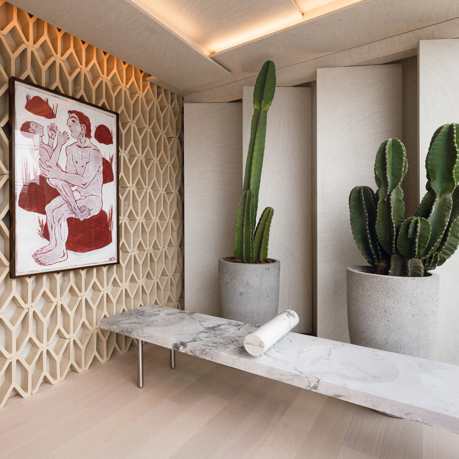 Sala com piso de madeira clara tauari, com um banco em mármore, dois vasos com cactos, uma parede com desenhos em relevo e um quadro com um desenho vermelho-escuro.